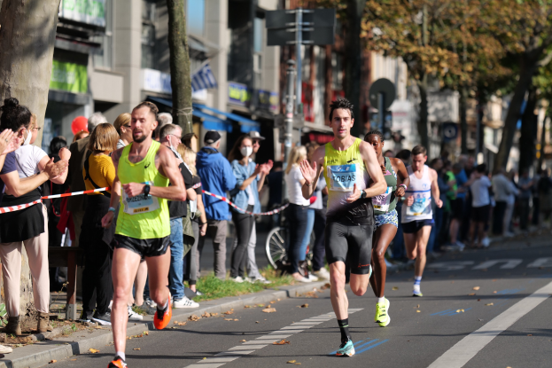 Gruppe von Läufern bei einem Marathon auf einer Stadtstraße mit Zuschauern auf der linken Seite, Bäumen, Gebäuden und einem klaren blauen Himmel im Hintergrund.