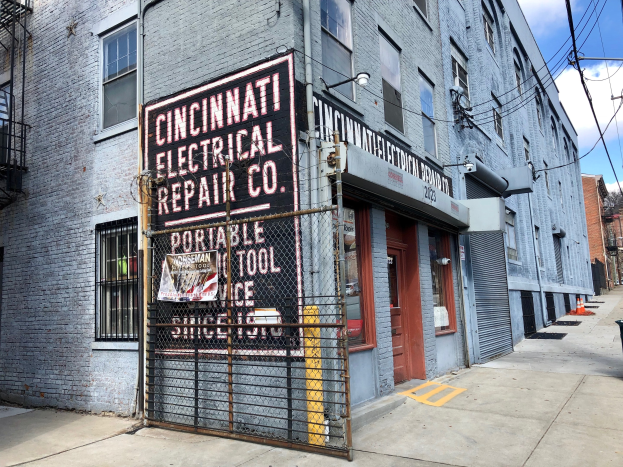 Außenansicht des Cincinnati Electrical Repair Co. Gebäudes mit einer Schautafel, einem Metallzaun, einem Müllbehälter, Polen, Drähten, Bäumen und einem bewölkten Himmel.