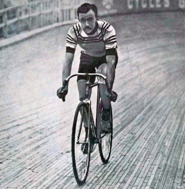 Schwarz-weiß-Foto eines Mannes, der auf einer Bahnradelt, mit einem Zaun und einer Tafel im Hintergrund.