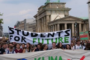 Eine Gruppe von Schülern marschiert in Berlin und hält ein buntes Banner mit der Aufschrift "Schüler für die Zukunft" in den Händen, während im Hintergrund Gebäude, Bäume und Himmel zu sehen sind.