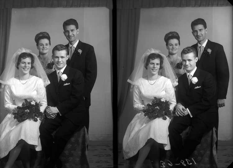 Ein Schwarz-Weiß-Foto eines Brautpaares, das für ein Hochzeitsfoto posiert, wobei die Braut in einem weißen Kleid und Schleier mit einem Strauß Blumen in der Hand steht, der Bräutigam hinter ihr steht und lächelt, und zwei weitere Personen hinter ihnen ebenfalls lächeln, mit einem Vorhang und einer Wand im Hintergrund.