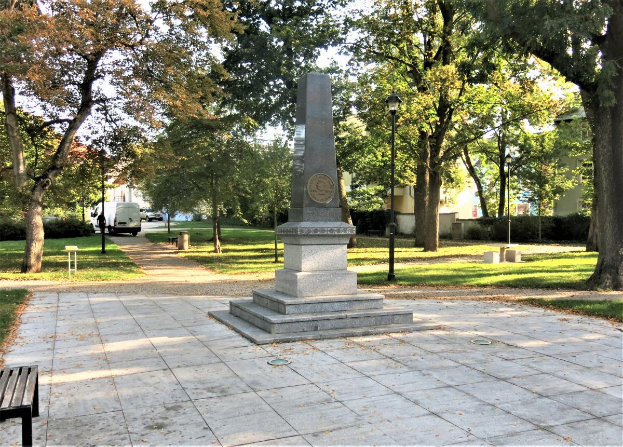 Ein Denkmal steht in einem Park mit einer Bank auf der linken Seite, umgeben von Bäumen, Laternen und Fahrzeugen auf einer nahen Straße, mit Gebäuden und einem klaren blauen Himmel im Hintergrund.