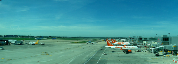 Eine Flughafen-Szene mit Flugzeugen, Gebäuden, Polen, Fahrzeugen, Bäumen, Himmel, Straße und einem Zaun im Vordergrund.