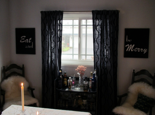 Ein Tisch mit Kerzen und Weinflaschen, ein Fenster mit einem Vorhang, Fotorahmen an der Wand und eine Gebäudeansicht draußen.