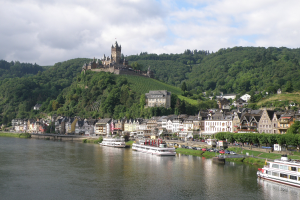 Ein malerischer Blick auf den Rhein in Deutschland mit einer Burg auf einem Hügel, Booten auf dem Fluss, Fahrzeugen auf einer näheren Straße und einer bewölkten Himmel.