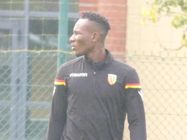 Ein Mann In Einer Schwarzen Jacke Steht Auf Einem Fussballfeld Umgeben Von Einem Zaun Und Einem Baum Und Sieht konzentriert Aus During A Training Session.