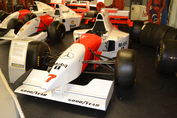 Eine Gruppe von Rennwagen in einem Museum ausgestellt, umgeben von Menschen, ein Schild mit Text, ein Seil und eine Wand im Hintergrund.
