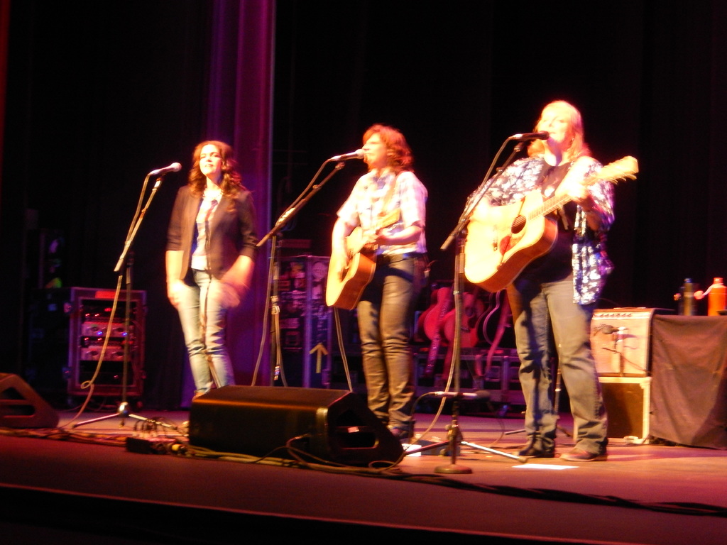 Drei Frauen performen auf der Bühne eines Musikkonzerts, singen in ein Mikrofon und spielen eine Gitarre, mit einem Tisch voller Wasserflaschen und zusätzlichen Gitarren dahinter.