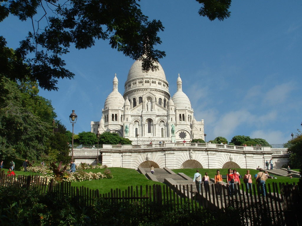 Ein weißes Gebäude, umgeben von einem Zaun, Gras, Bäumen und Menschen, mit Pflanzen und Pfählen in der Nähe.
