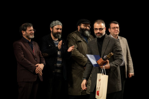 Eine Gruppe von Mönnen steht auf einer Bühne, einer hölt eine Tasche und eine Trophî, mit einem dunklen Hintergrund, der die Eröffnung der Berliner Filmfestspiele suggeriert.