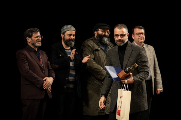 Eine Gruppe von Mönnen steht auf einer Bühne, einer hölt eine Tasche und eine Trophî, mit einem dunklen Hintergrund, der die Eröffnung der Berliner Filmfestspiele suggeriert.