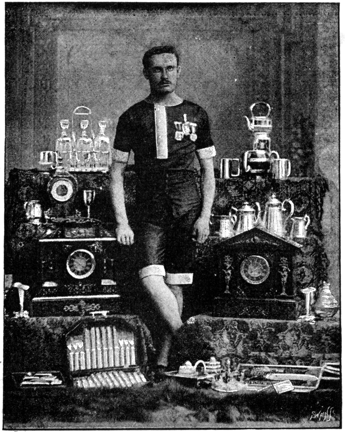 Schwarzes und weißes Foto eines Mannes in einem Fußballtrikot, der vor einem Tisch mit Pokale und Trophäen steht, die mit '1909-1910 WM-Sieger' beschriftet sind.