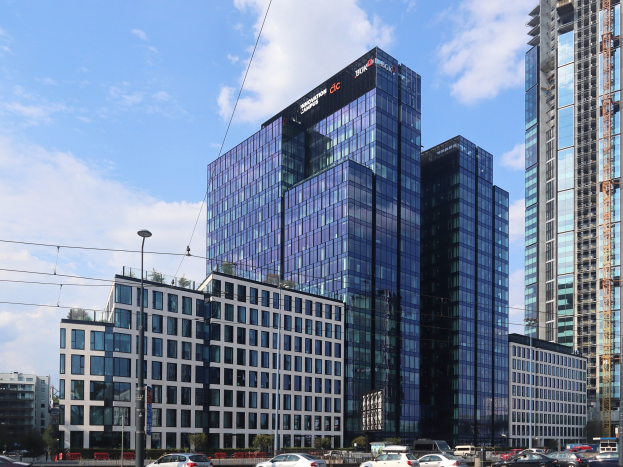 Großes modernes Bürogebäude mit vielen Fenstern, die Deutsche Bank Hauptverwaltung in Frankfurt, Deutschland, umgeben von städtischer Infrastruktur einschließlich Straßenlaternen, Fahrzeugen, Grünflächen und einem bewölkten Himmel.