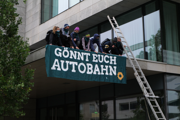 Menschen auf einem Gebäude stehend, die eine Fahne halten, mit einem Baum links und Glasfenstern im Hintergrund, wahrscheinlich protestierend in Unterstützung der deutschen Regierung.