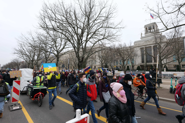 Eine große Gruppe von Menschen marschiert auf einer Straße in Washington, D.C., mit Schildern und Bannern, einige fahren Fahrräder, unter einem klaren blauen Himmel.