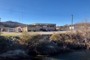 Ein Fluss fließt durch ein grünes Feld neben einem Gebäude, mit Bäumen, Pflanzen, Gras, Steinen, Straßenmasten, Straßenlaternen, Strommasten, Stromkabeln und Hügeln unter einem klaren blauen Himmel an der Stelle einer geplanten Wasseraufbereitungsanlage.