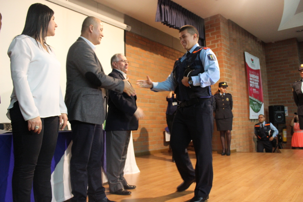Eine Gruppe von Menschen auf einer B√ľhne, einer in Uniform mit einer M√ľtze, eine Frau in einem wei√üem Hemd, mit einem Banner, Lautsprecher, Vorh√§ngen, Tisch und Wandmonitor im Hintergrund.