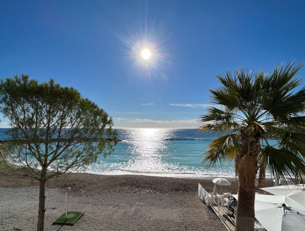 Ein sonniger Strand mit Palmen, Sonnenschirmen und üppiger Vegetation, vor einem strahlend blauen Himmel mit der Sonne im Hintergrund.