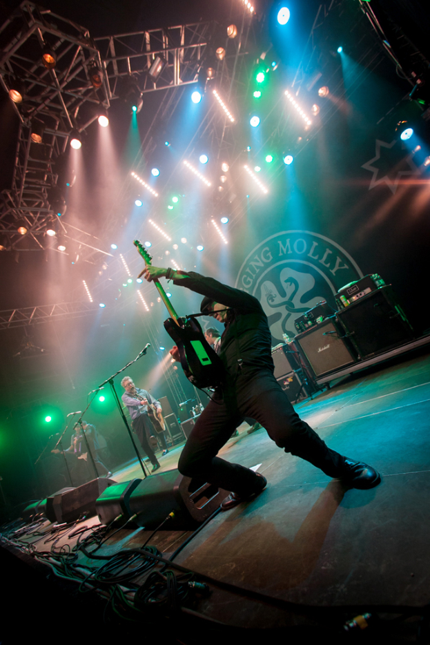 Ein musikalisches Konzert mit drei Künstlern auf der Bühne: zwei Gitarristen rechts mit Mikrofonen davor, ein Hintergrund mit dem Wort "MOLLEY" und zwei Schlangensymbolen sowie Scheinwerfer darüber.