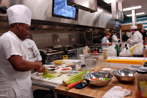 Eine Gruppe von Köchen in einer Küche beim Zubereiten von Speisen, mit einem Koch im Vordergrund in einer weißen Schürze und Mütze, der ein Messer hält und von verschiedenen Utensilien, Schüsseln, Gemüse, einem Handy, Flaschen und anderen Gegenständen umgeben ist, während im Hintergrund ein Fernseher und Deckenlampen zu sehen sind.