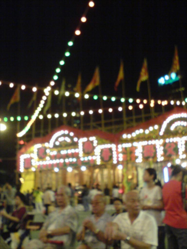 Menschen auf Stühlen im unteren Bildbereich, dekorative Lichter in der Mitte und Flaggen oben unter einem nächtlichen Himmel.