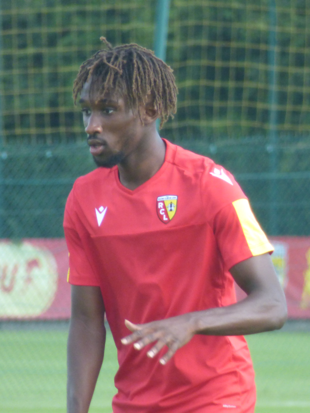 Ein Mann mit Dreadlocks steht auf einem Fußballfeld und trägt ein rotes T-Shirt und Shorts, im Hintergrund gibt es ein Netz und eine Tafel mit der Aufschrift "RCCL - Ghanaian Premier League".