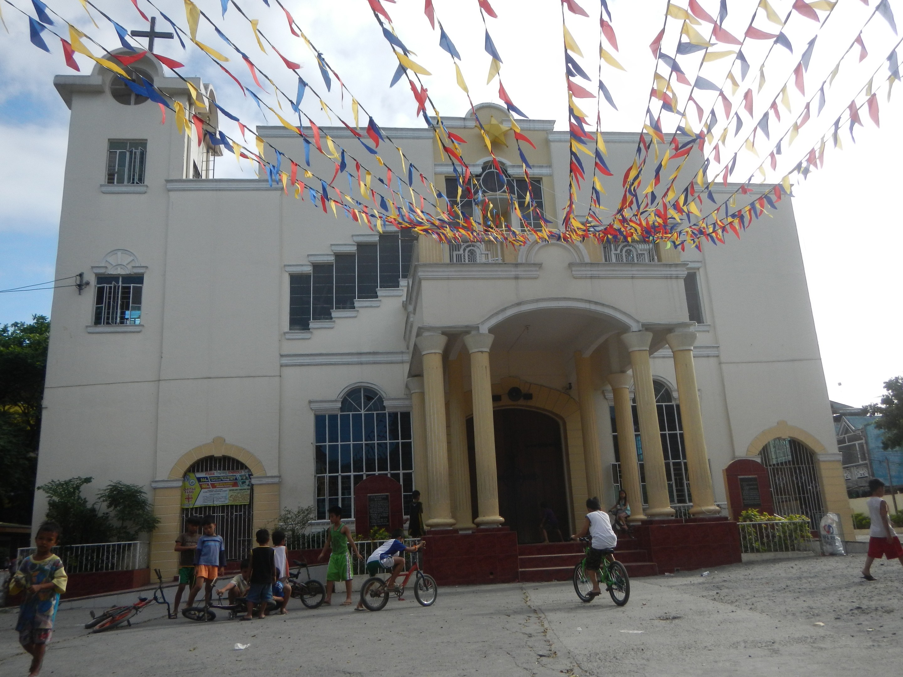 Eine Gruppe von Menschen, die auf Fahrrädern vor einer weißen Kirche mit architektonischen Details, Pflanzen, einem Zaun, einer Fahne, Bäumen, einem bewölkten Himmel und bunten Fahnen fährt.