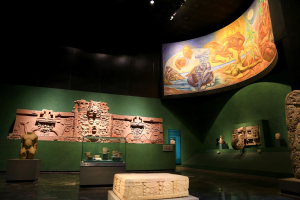 Ein Museumsraum mit verschiedenen Artefakten, darunter Skulpturen, Tischgegenstände, eine Texttafel, eine Wandmalerei und Deckenleuchten im Mexiko-Stadt-Museum der Anthropologie.