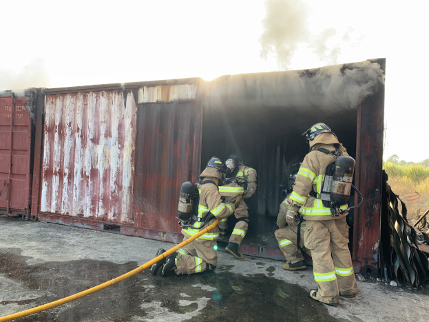 Feuerwehrleute in Schutzausrüstung löschen ein Feuer in einem Seecontainer, wobei Rauch aufsteigt und Grünfläche im Hintergrund zu sehen ist.
