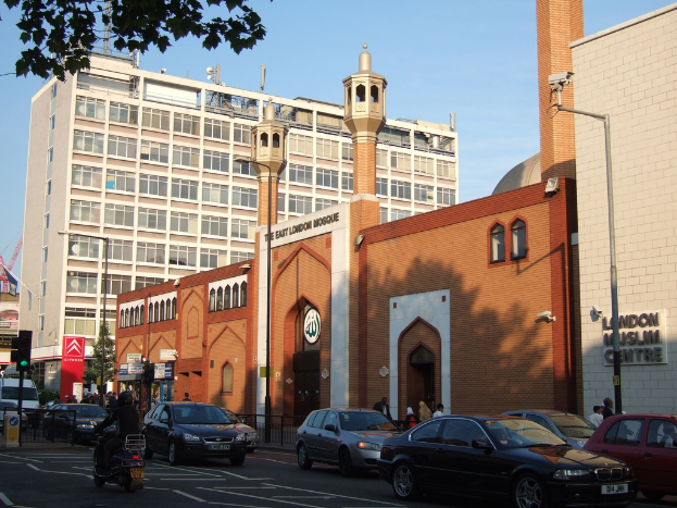 Ein Moschee-Gebäude mit Fenstern und Glastüren, das eine Namensplakette an der Wand, Straßenlaternen, eine Ampel an einem Mast, Fahrzeuge auf der Straße, Gruppen von Menschen, einen Baum und eine Überwachungskamera an einem Mast zeigt.