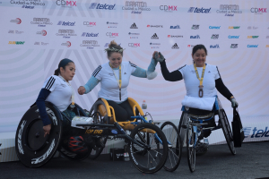 Drei Frauen im Rollstuhl auf einer Rennbahn, jede trägt eine Medaille, mit einem Banner im Hintergrund.
