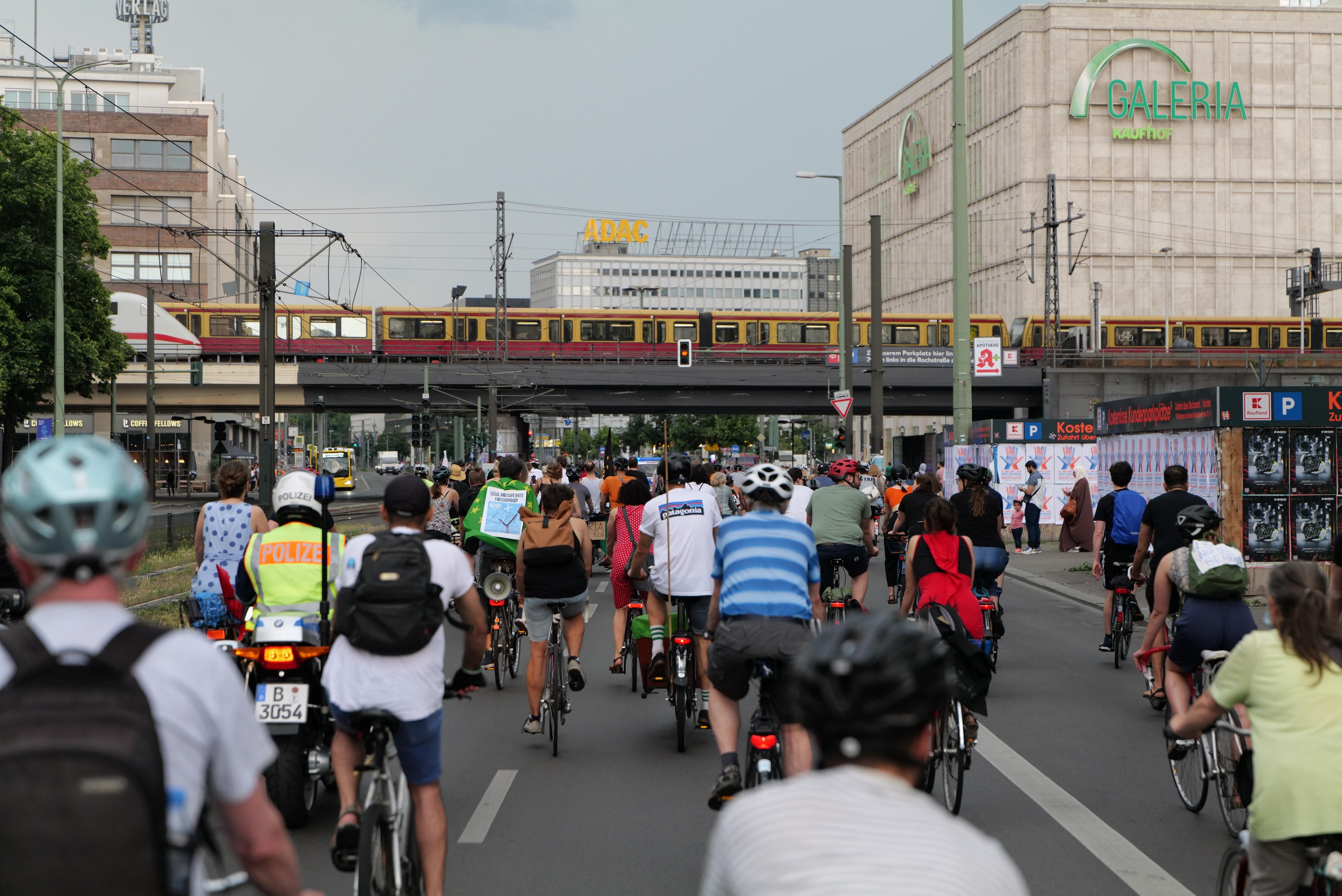 Eine Gruppe von Menschen, die auf Fahrrädern eine Straße entlangfahren, die von hohen Gebäuden gesäumt ist, einige tragen Helme und Taschen, mit einem Zug auf einem Bahngleis, Strommasten, Bäumen und einem klaren blauen Himmel im Hintergrund.