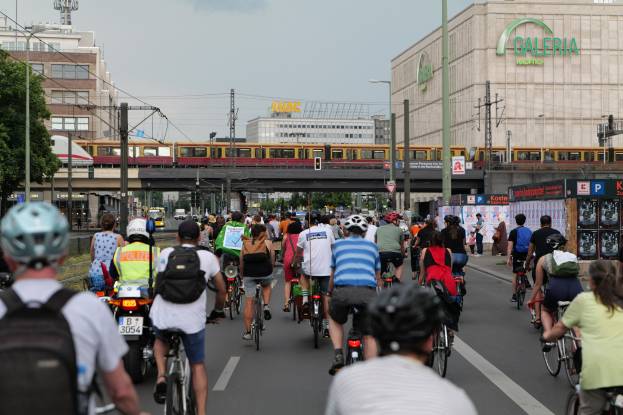 Eine Gruppe von Menschen, die auf Fahrrädern eine Straße entlangfahren, die von hohen Gebäuden gesäumt ist, einige tragen Helme und Taschen, mit einem Zug auf einem Bahngleis, Strommasten, Bäumen und einem klaren blauen Himmel im Hintergrund.