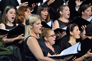 Eine Gruppe von Frauen, die in einem Chor singen, einige halten Bücher in den Händen, mit einer Person, die im Vordergrund sitzt und einem unscharfen Hintergrund.