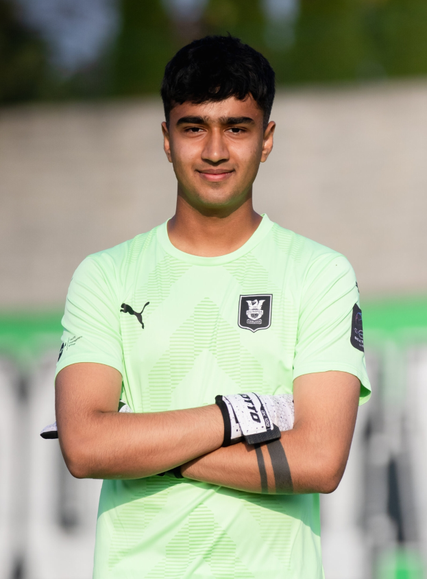 Ein junger Mann in einem grünen T-Shirt steht auf einem Fußballfeld und schaut mit verschränkten Armen und einem Lächeln in die Kamera, mit einem unscharfen Hintergrund.