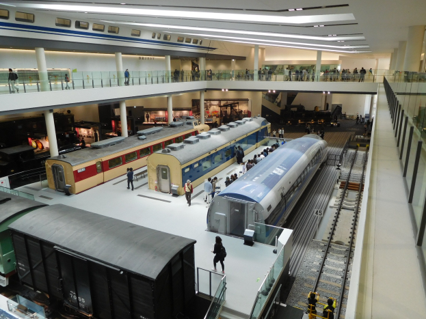 Ein großer Museumsraum mit verschiedenen Zügen in unterschiedlichen Größen und Farben, mit Menschen, die sie vom Boden, Geländern und Säulen unter Deckenlampen beobachten.