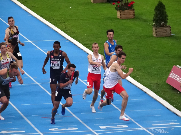Eine Gruppe von Männern, die auf einer blauen Bahn laufen, umgeben von grünem Gras und Blumentöpfen mit einer Texttafel auf der rechten Seite, die auf einen Wettbewerb hinweist.