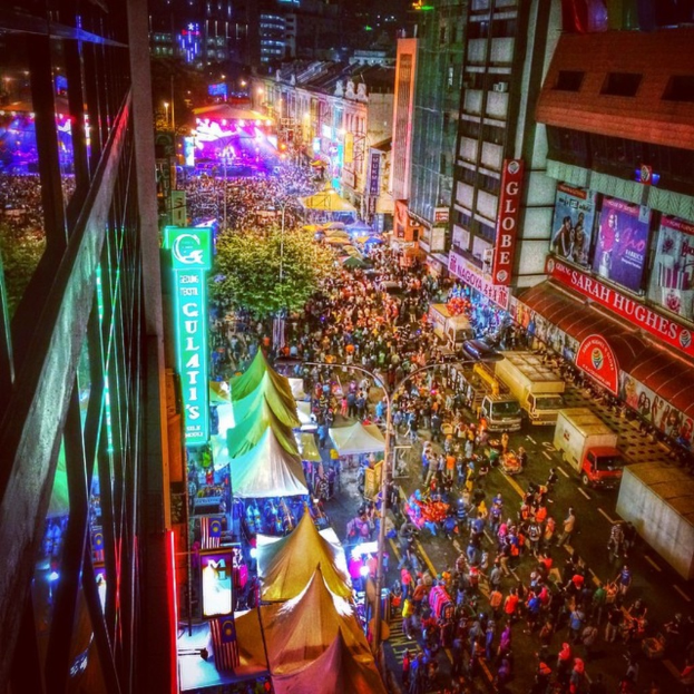 Eine lebhafte Karnevalszenen mit einer großen Menge auf einer Straße, umgeben von Zelten, Geschäften, Gebäuden und Bannern, mit einem hell erleuchteten Bereich im Hintergrund.