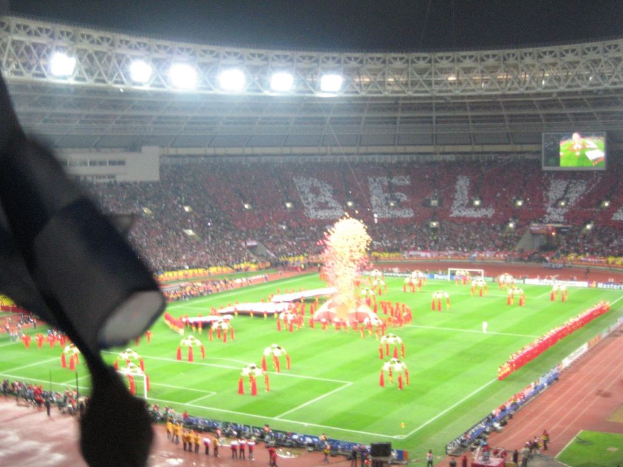 Ein Stadion voller Zuschauer, mit Flutlicht, Eisengittern, Werbetafeln, Anzeigetafeln, Sicherheitskameras und dekorativen Fahnen.