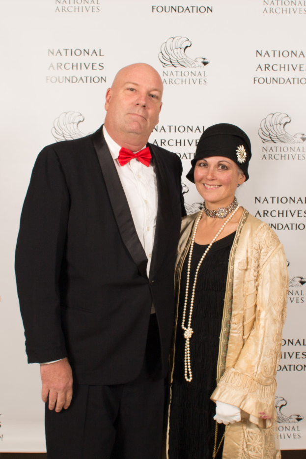 Ein Mann in einem schwarzen Anzug mit einem weißen Hemd und einer schwarzen Fliege steht neben einer Frau in einem eleganten Kleid mit einem gemusterten Schal auf einem roten Teppich, mit einem weißen Banner mit Text im Hintergrund.