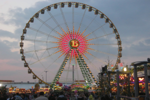Ein mit bunten Lichtern beleuchtetes Riesenrad steht inmitten einer Menschenmenge auf einem Vergnügungspark, umgeben von Pfählen, Bäumen und anderen Gegenständen, mit einem Himmel voller weißer, flauschiger Wolken im Hintergrund.