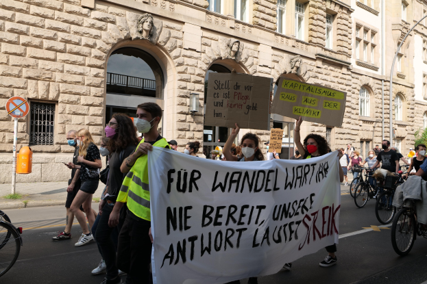 Eine Gruppe von Menschen marschiert auf einer Demonstration gegen die Anti-Kriegsbewegung in Berlin, einige halten Schilder und andere fahren Fahrräder, mit einem Gebäude im Hintergrund, das Fenster, Bögen, Säulen und Skulpturen aufweist.