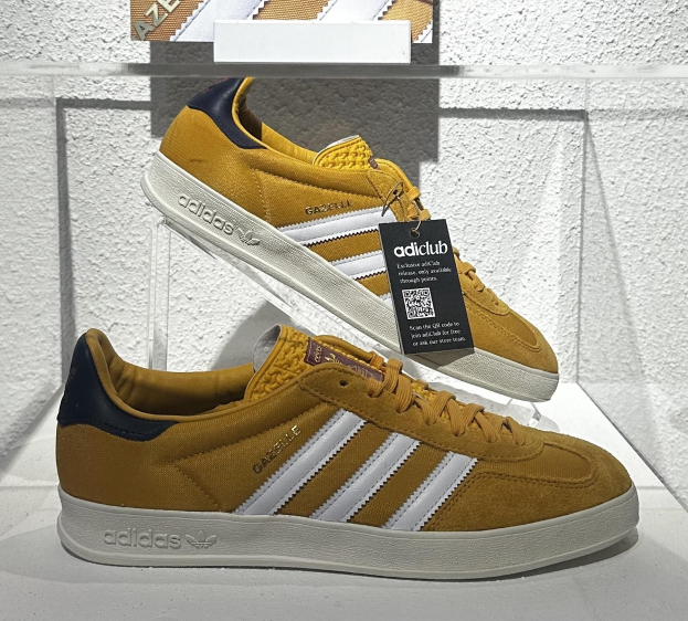 Ein Paar gelbe, weiße und navyblaue adidas Gazelle OG-Schuhe mit einem Anhänger, auf einer weißen Oberfläche gegen eine weiße Wand und ein Glasregal gestellt.