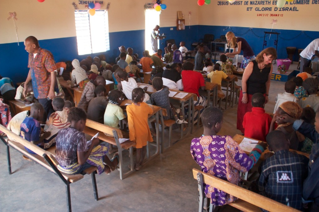 Kinder auf Bänken in der Vordergrund, mit Erwachsenen, die sie führen, und einem Hintergrund mit Stühlen, einer Tür, einem Fenster, einem Tisch, Ballons und Text oben.