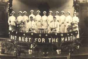 Eine Gruppe von Frauen in weißen Kleidern und Hauben steht auf einer Bühne mit einem Banner, auf dem 'Unser Bestes für den Meister' steht, umgeben von Blumentöpfen und Türen und einer Wand im Hintergrund.