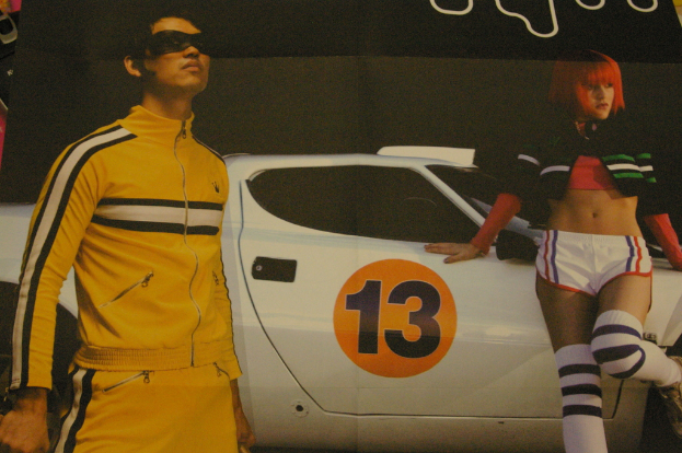 Ein Mann in einer gelben Jacke und einem Trainingsanzug posiert vor einem weißen Rennwagen, neben ihm eine Frau in einer grünen Jacke, vor einem schwarzen Hintergrund.