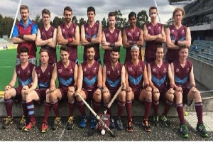 Eine Gruppe von Männern, die auf einem Feld mit Hockey-Schlägern feiern, Bäume und ein Stadion mit Zuschauern im Hintergrund.