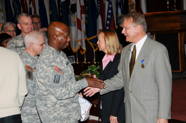 Eine Gruppe von Menschen mit zwei Personen, die sich die Hände schütteln, im Hintergrund Fahnen an Stangen, ein Podium mit einem Mikrofon und in der Nähe eine Topfpflanze.