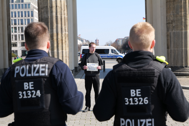 Drei Polizeibeamte stehen vor einem Gebäude, wobei ein Mann in der Mitte ein Papier hält, flankiert von Säulen, mit Fahrzeugen, Menschen, Bäumen, Gebäuden und dem Himmel im Hintergrund.
