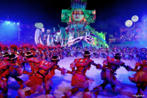 Eine Gruppe von Menschen in bunten Kostümen tanzt auf einer Bühne vor einem sitzenden Publikum, mit Lichtern, Pfählen, Ballons und einem Bildschirm, der eine Person in der Hintergrund zeigt, unter einem sichtbaren Himmel, bei der Rio-Karneval 2014.
